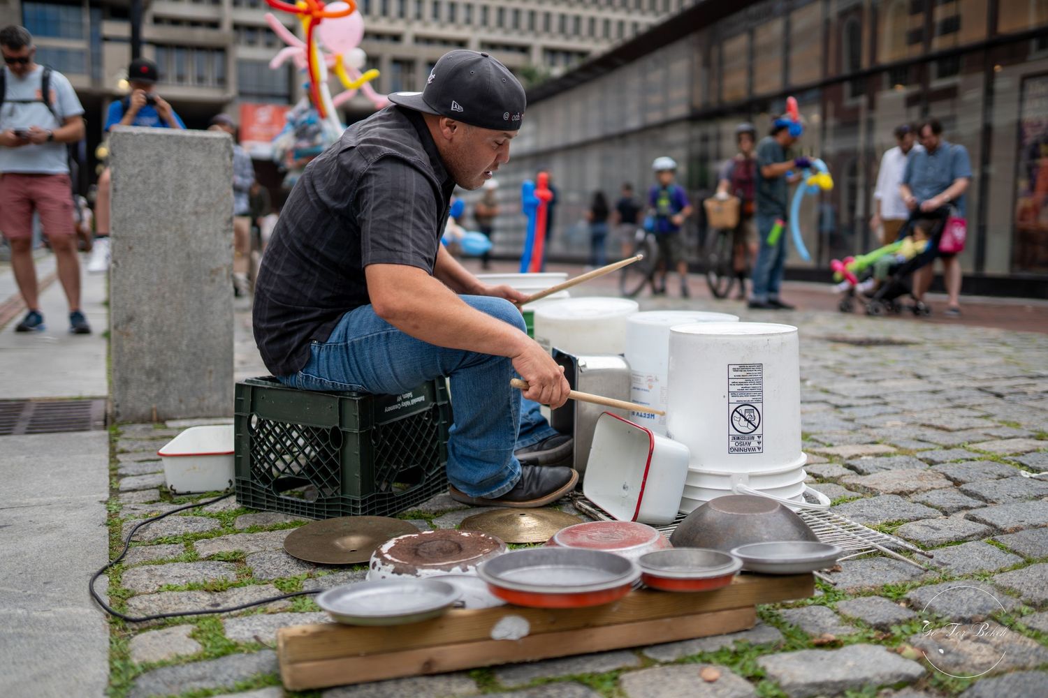 Street performer playing improvised drum kit made of buckets and pans at outdoor urban festival.