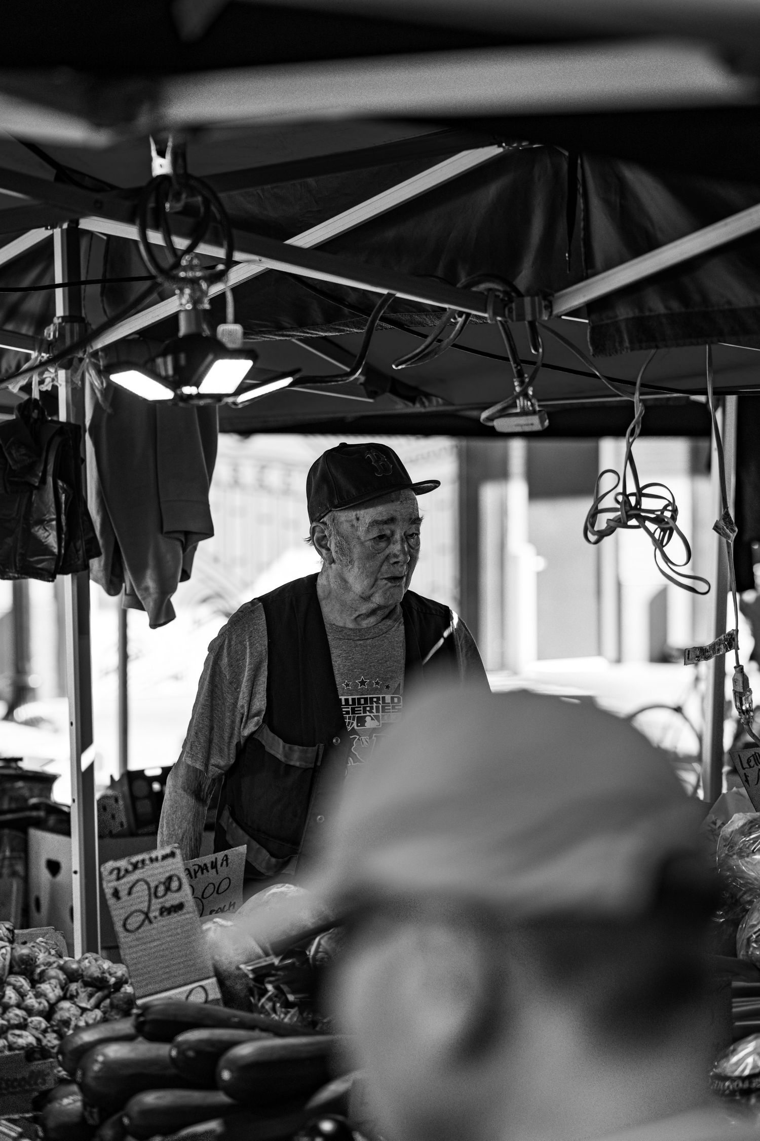 Black and white portrait of an elderly market vendor working under a canopy at an outdoor market.
