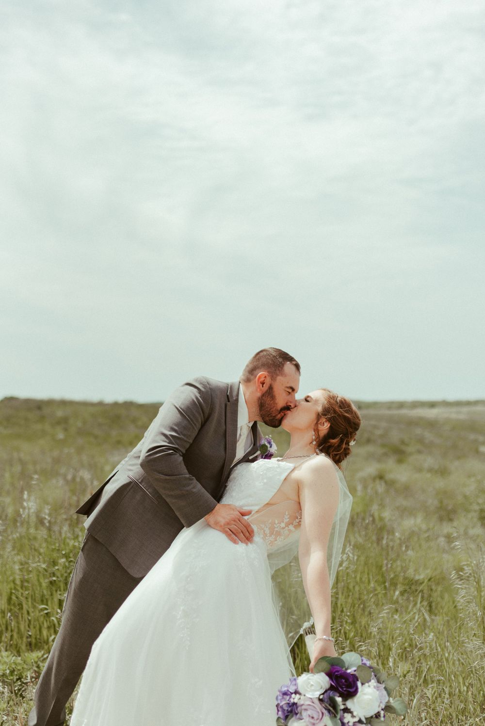 Intimate wedding portrait of couple kissing in a rustic outdoor setting with purple bouquet.