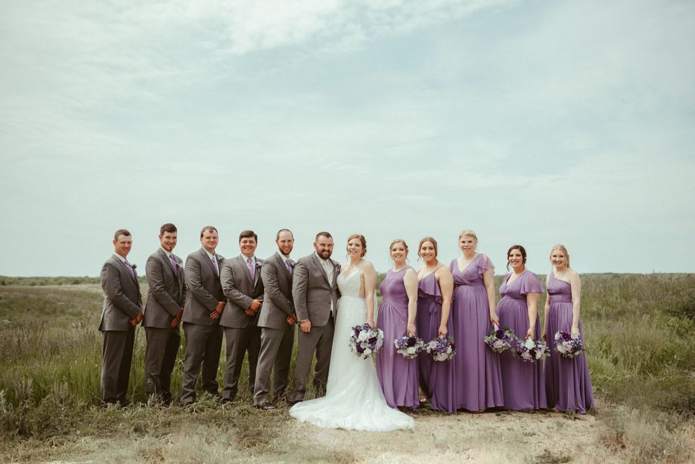 A wedding party poses together in a field with bridesmaids in purple dresses.