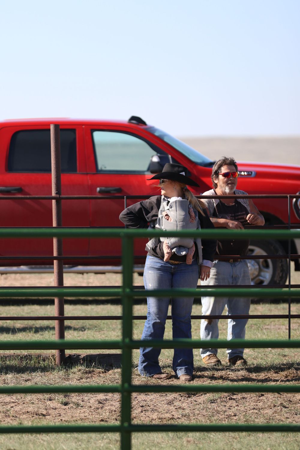 Red pickup truck parked behind green metal corral fencing at a ranch.