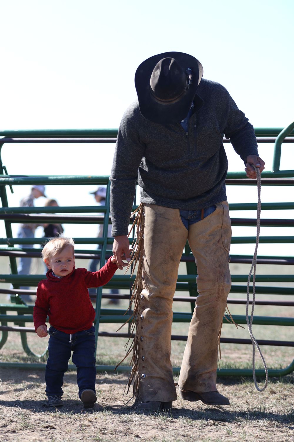 Cowboy in western attire with chaps and hat standing next to small child at rodeo.