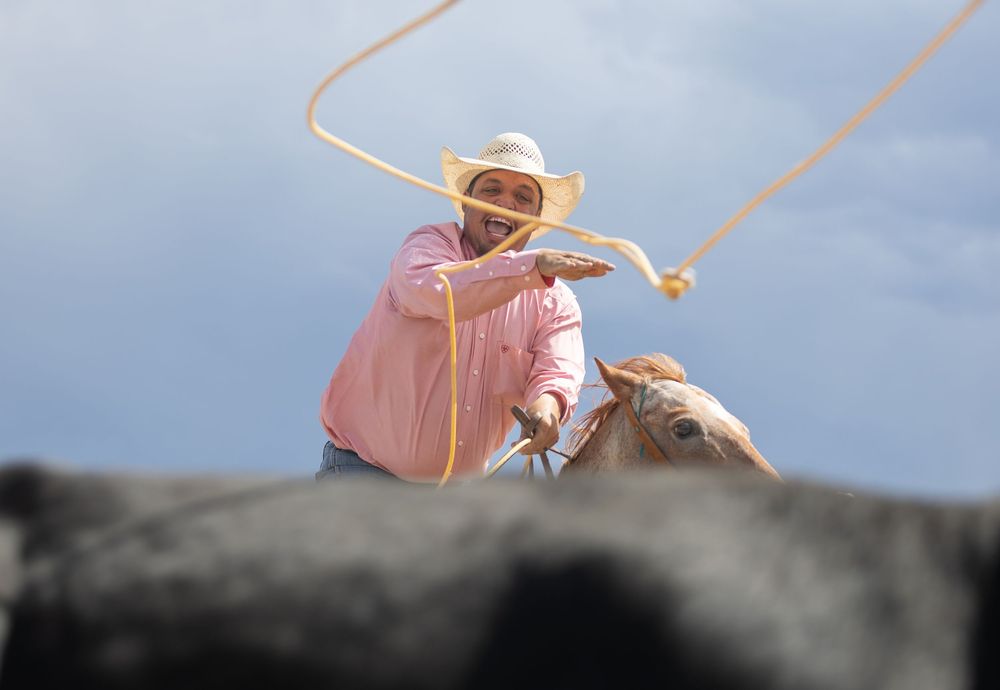 A cowboy in pink shirt and white hat swings a lasso against a cloudy sky background.