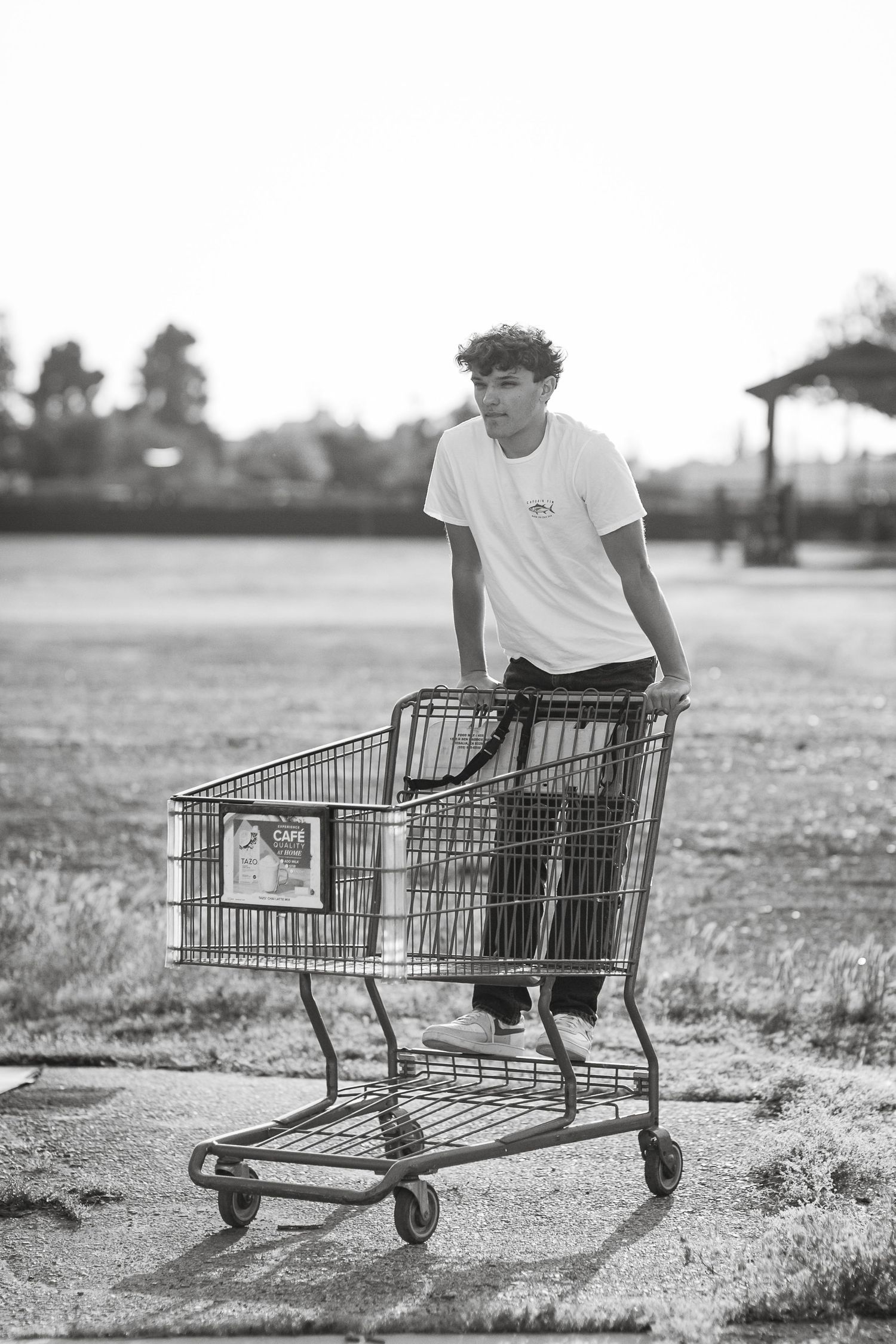 A person in a white t-shirt performs tricks while sitting in an empty shopping cart in a grassy field.