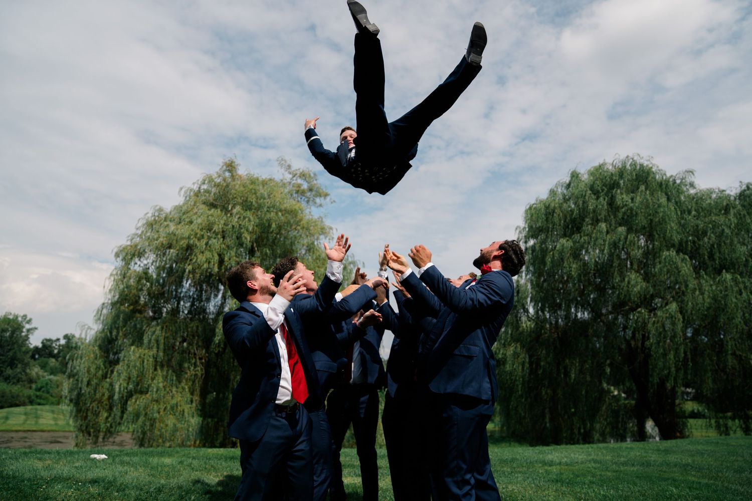 Wedding party in formal attire performs celebratory jumps on grassy lawn with willow trees.