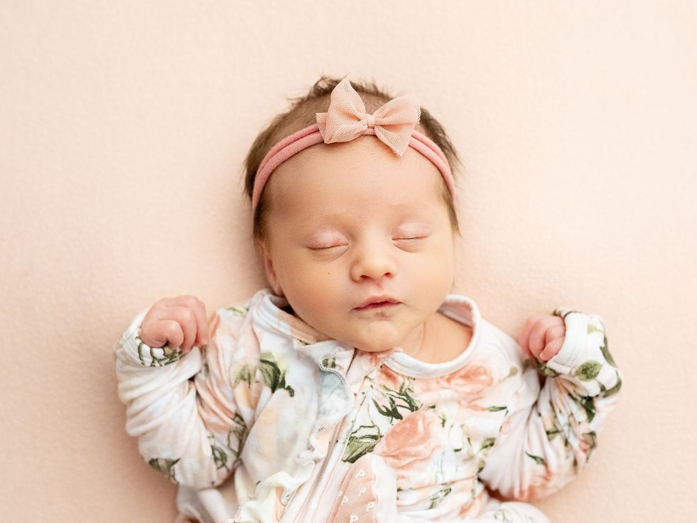 Sleeping baby in floral onesie and pink bow headband resting on beige backdrop for newborn photoshoot.