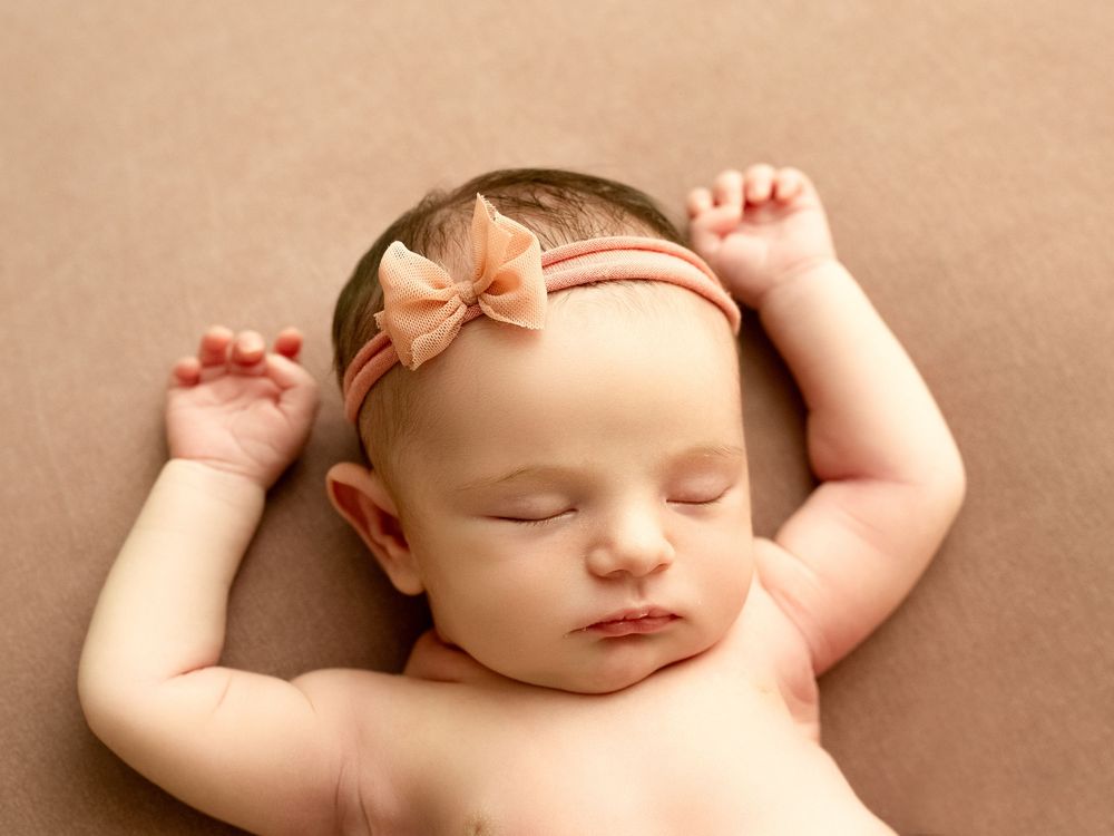 Peaceful sleeping newborn wearing a peach bow headband on soft brown backdrop.