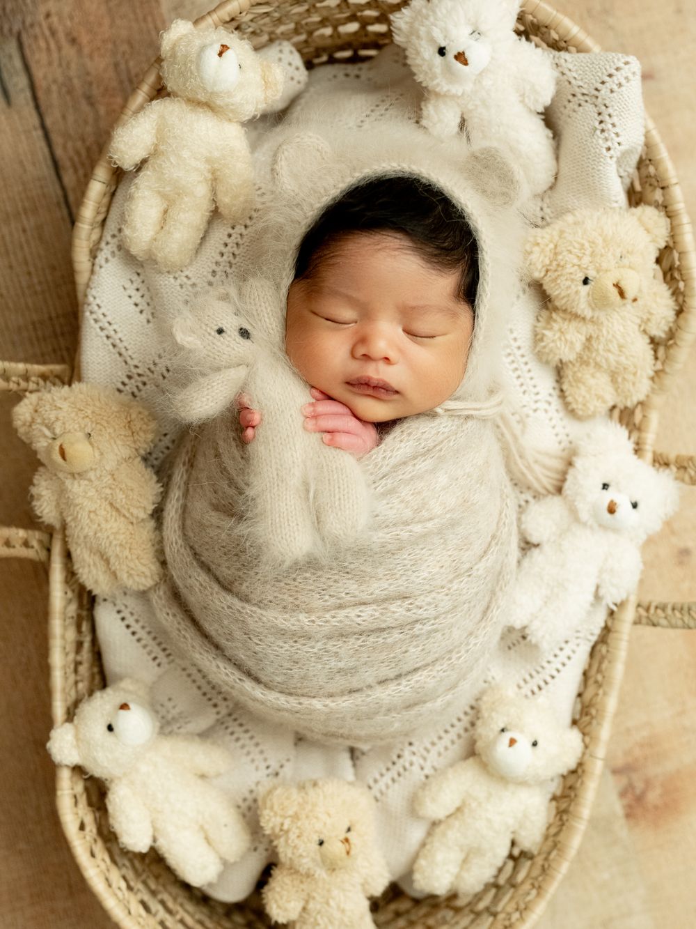 A sleeping newborn wrapped in soft white fabric surrounded by plush white teddy bears and knit textures.