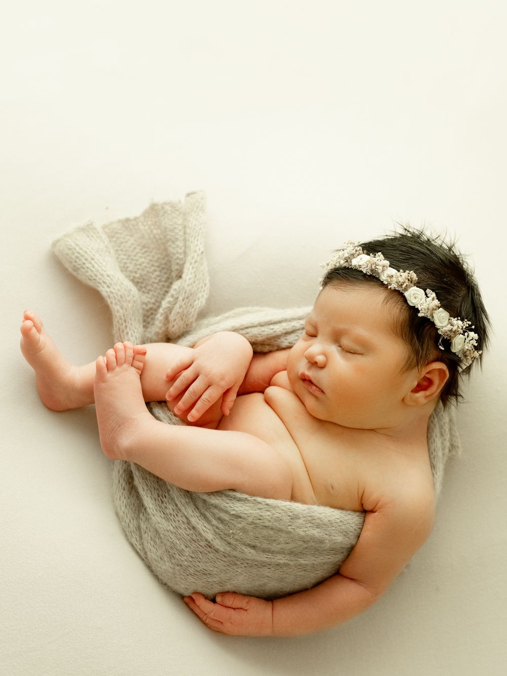 A sleeping newborn wrapped in soft gray fabric with a delicate floral headband, captured in a series of gentle poses.