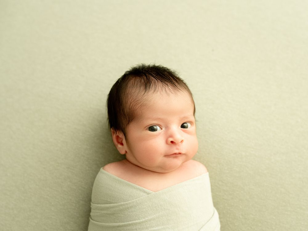 A baby sits against a plain wall wearing a white onesie, showing several playful expressions.