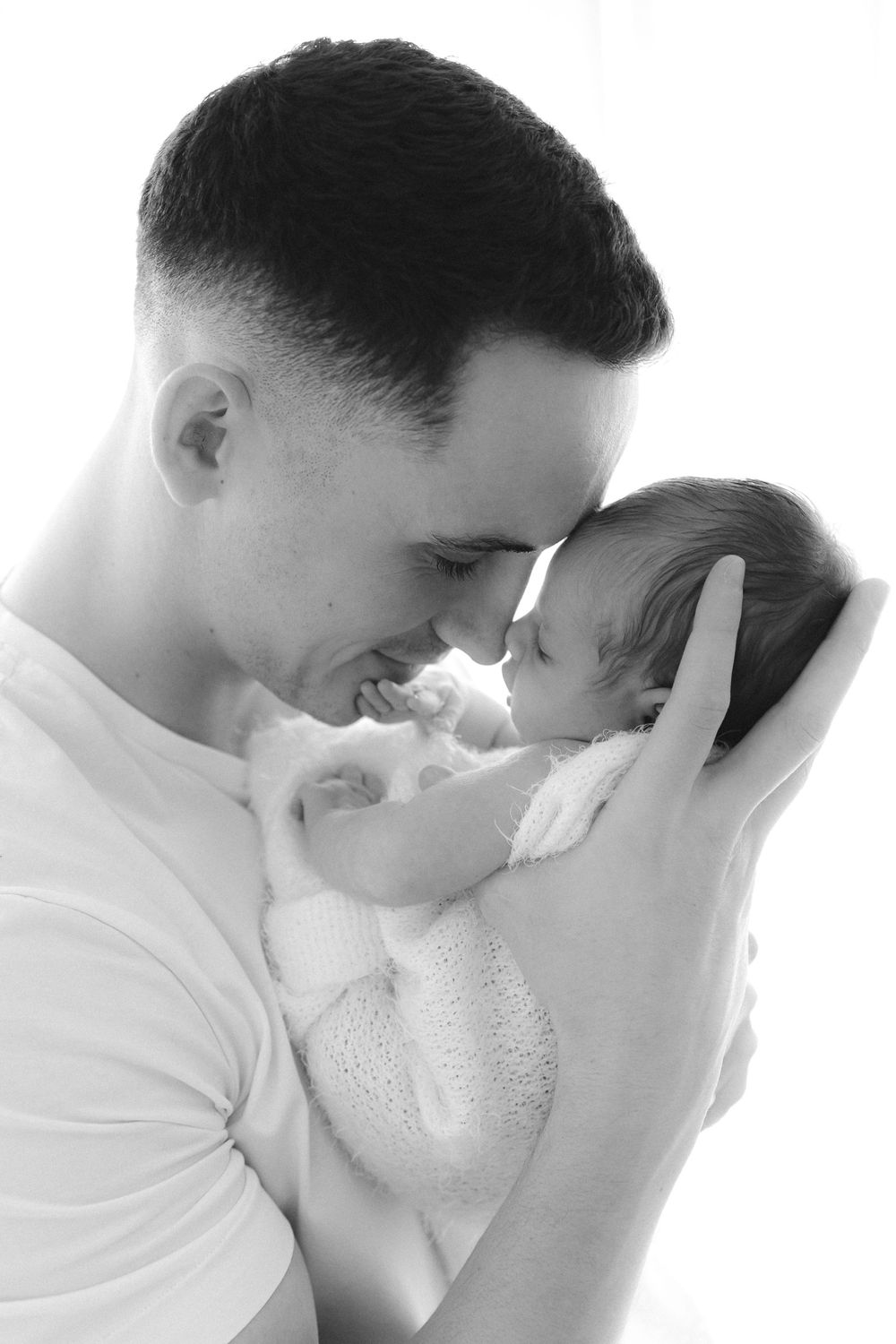 Black and white photo of a young father tenderly holding his newborn baby close, touching foreheads gently.