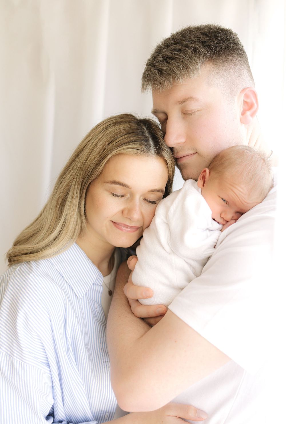 A couple tenderly holds their sleeping newborn baby, eyes closed, in a soft, bright studio setting.