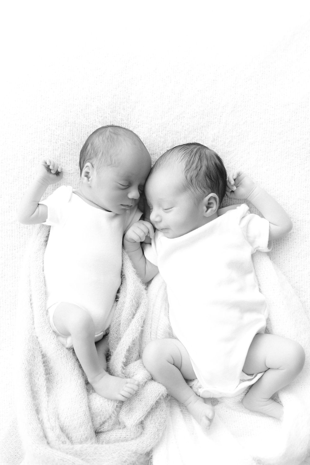 Two sleeping newborn twins lying side by side on a soft white blanket in a black and white photo.