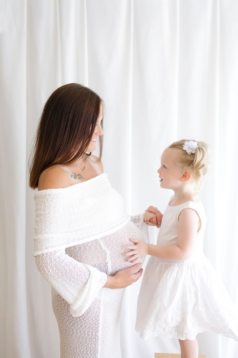 Pregnant woman in white lace dress with toddler girl in white dress touching baby bump against white curtains.