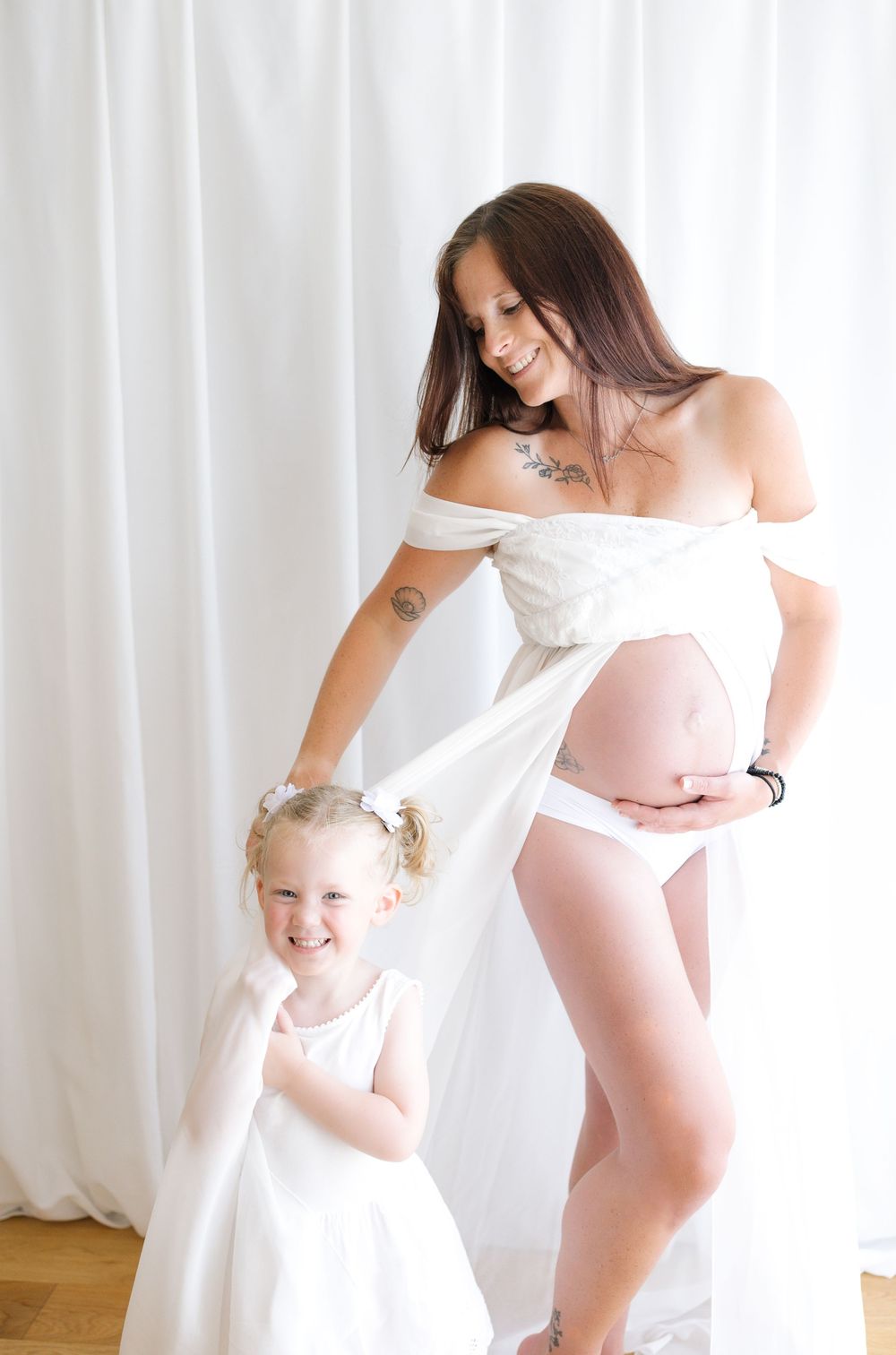 Pregnant woman in white wrap smiles at young girl in white dress during a bright maternity photo session.