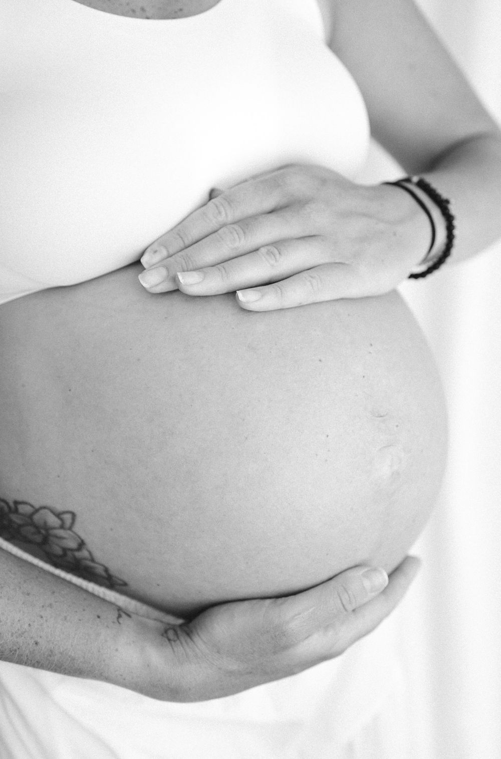 Black and white photo of a pregnant woman cradling her bare baby bump with both hands, wearing a bracelet.