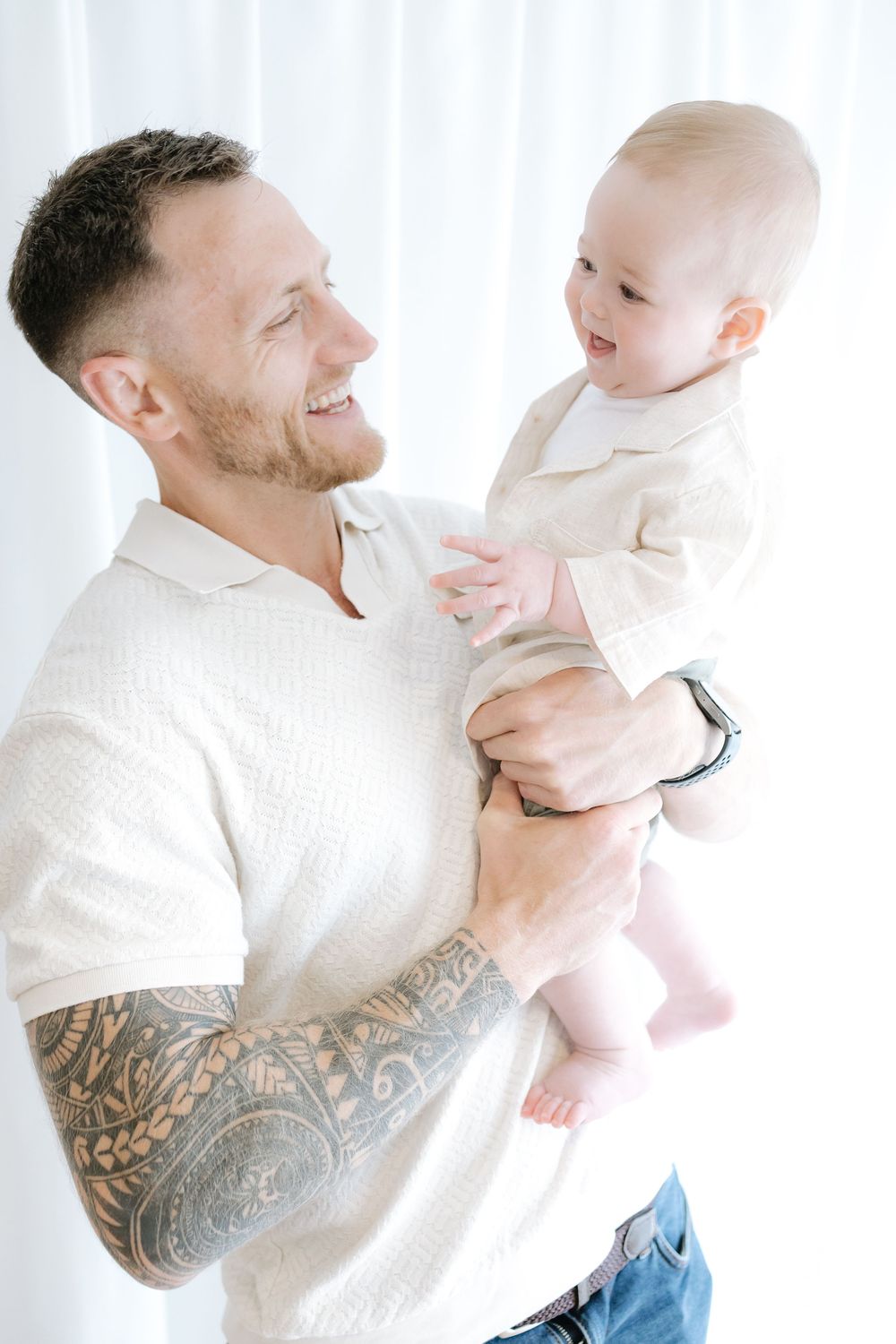Smiling tattooed father holding his happy baby in a bright white room.