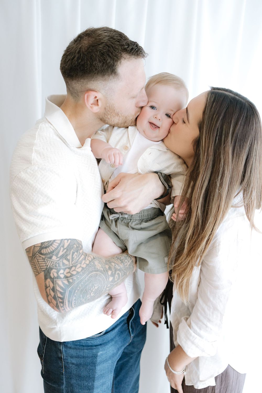 A young couple kisses their smiling baby on the cheeks in a bright, airy studio family portrait.