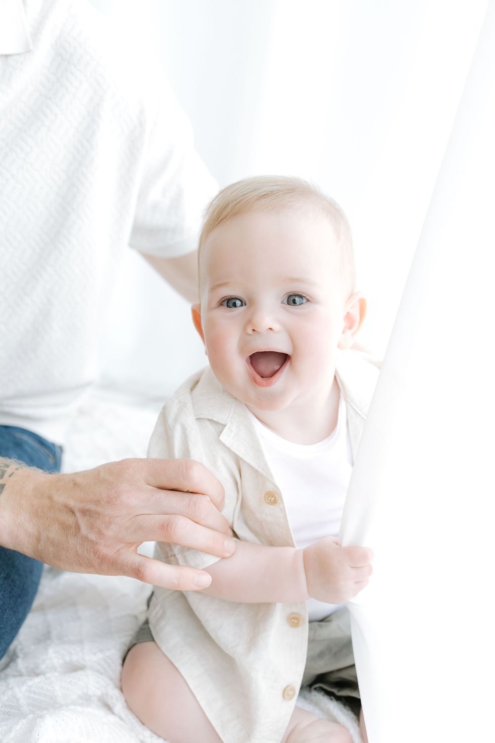 Happy laughing baby in beige outfit sitting with adult hands supporting them against white background.