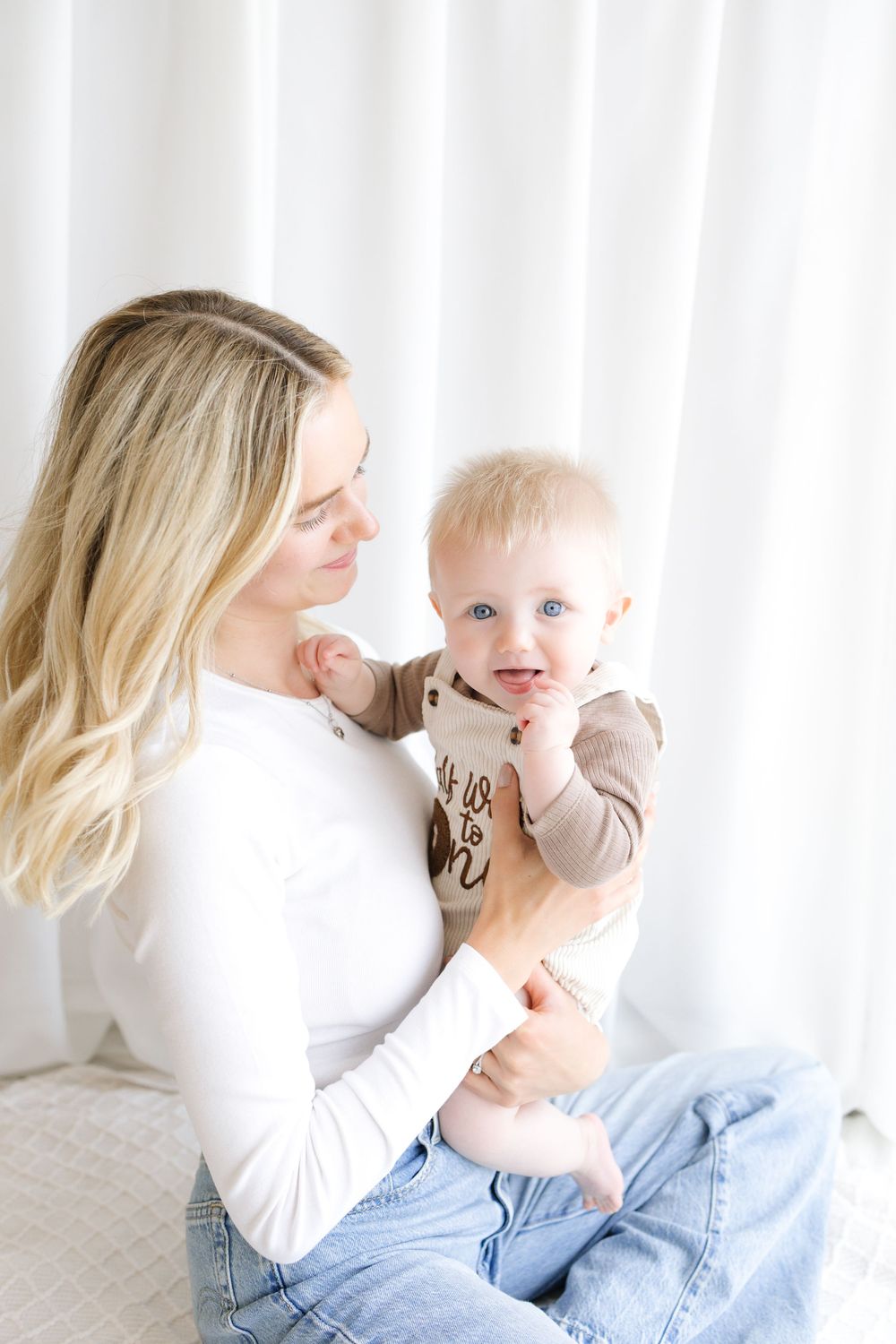 A young blonde mother in a white top and jeans holds her blue-eyed baby in a bright, airy white room.