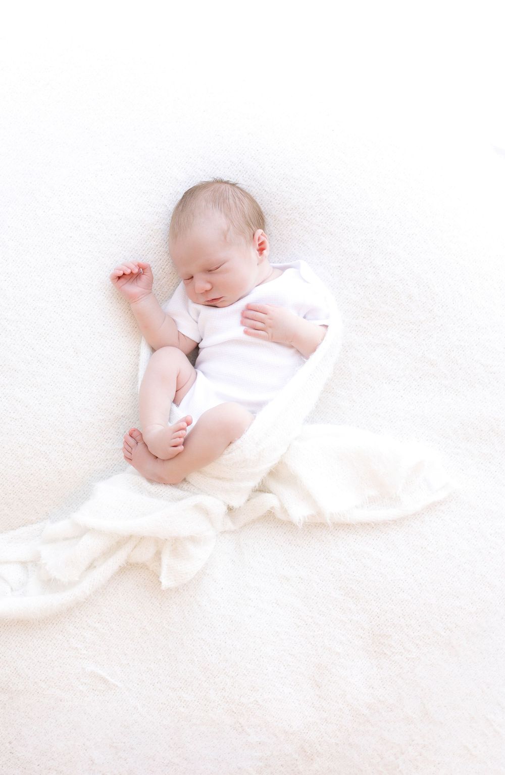 A peaceful newborn baby sleeping on a soft white blanket, dressed in a white onesie with tiny feet curled up.