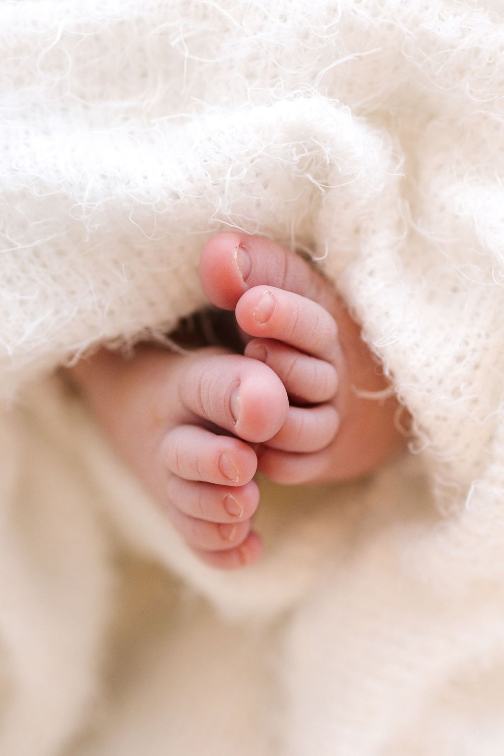 Close-up of tiny newborn baby toes peeking out from a soft white knit blanket.