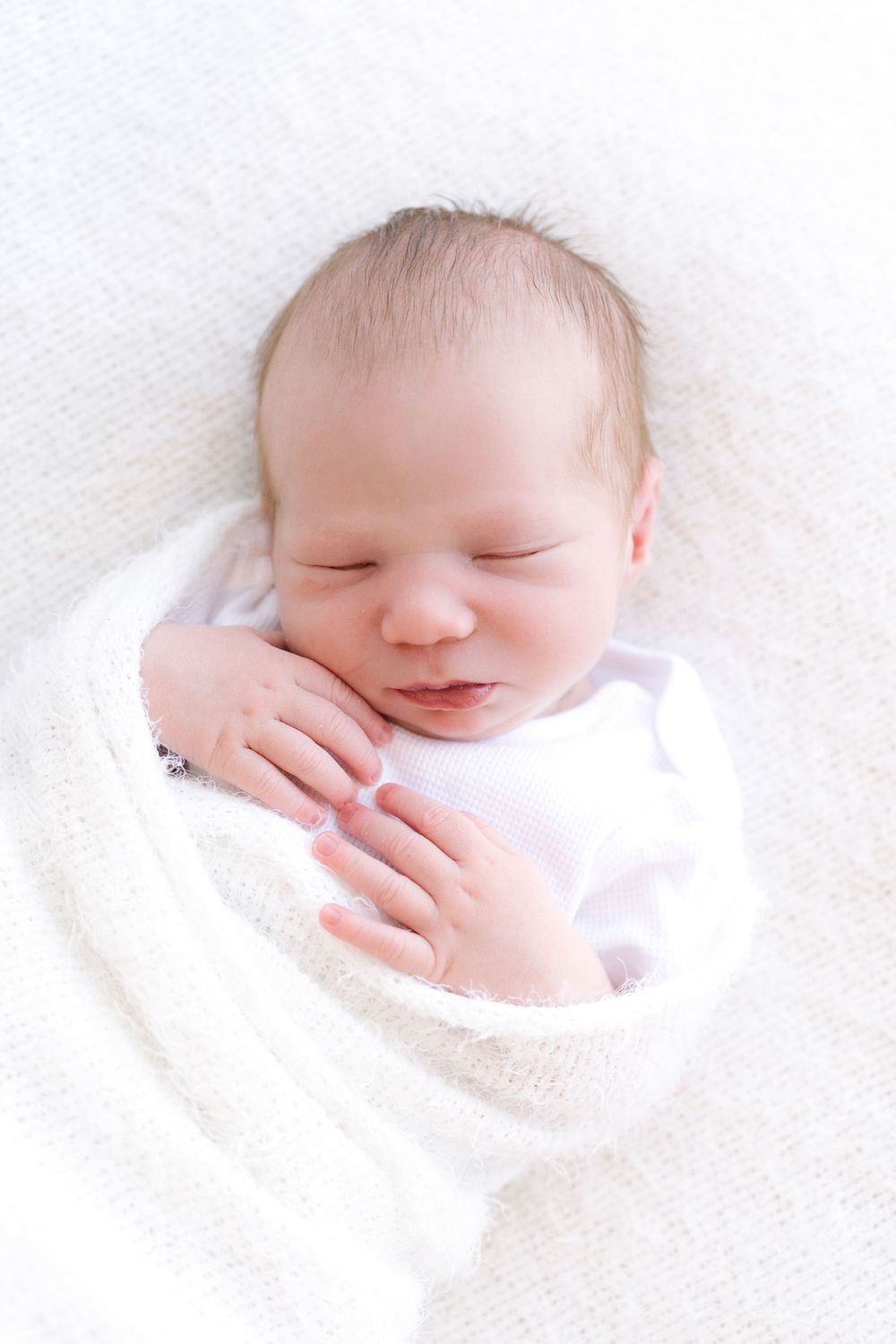 Peaceful newborn baby swaddled in white blanket sleeping with tiny hands visible near face.