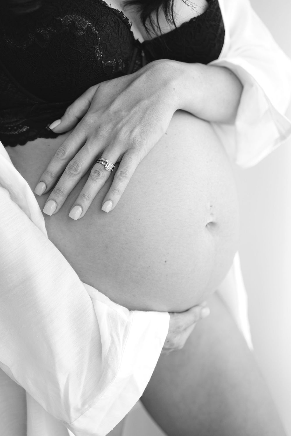 Black and white photo of a pregnant woman cradling her bare baby bump, wearing a black lace bra and open white shirt.