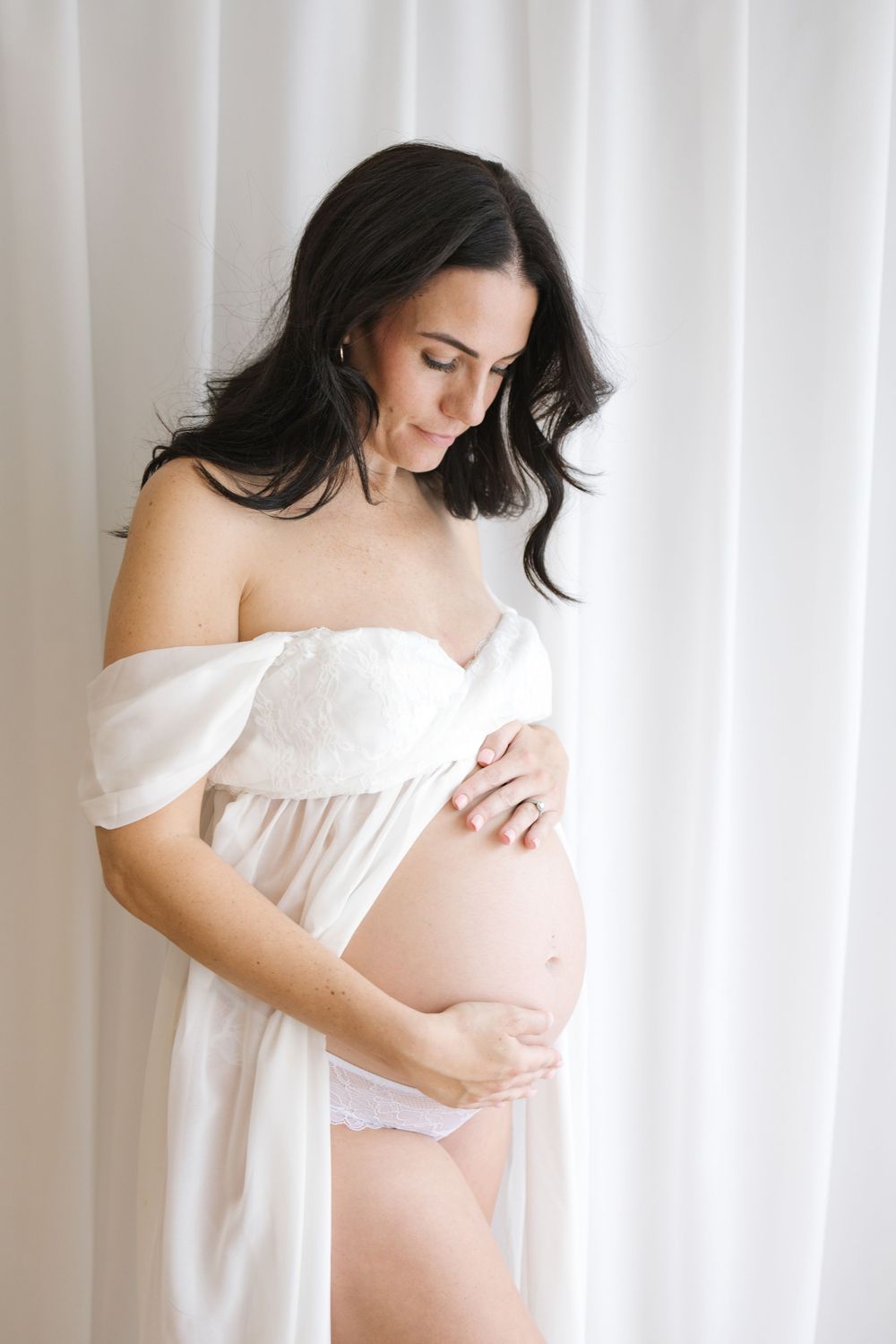 Pregnant woman in white lingerie and sheer robe cradling her baby bump, posing against soft white curtains.