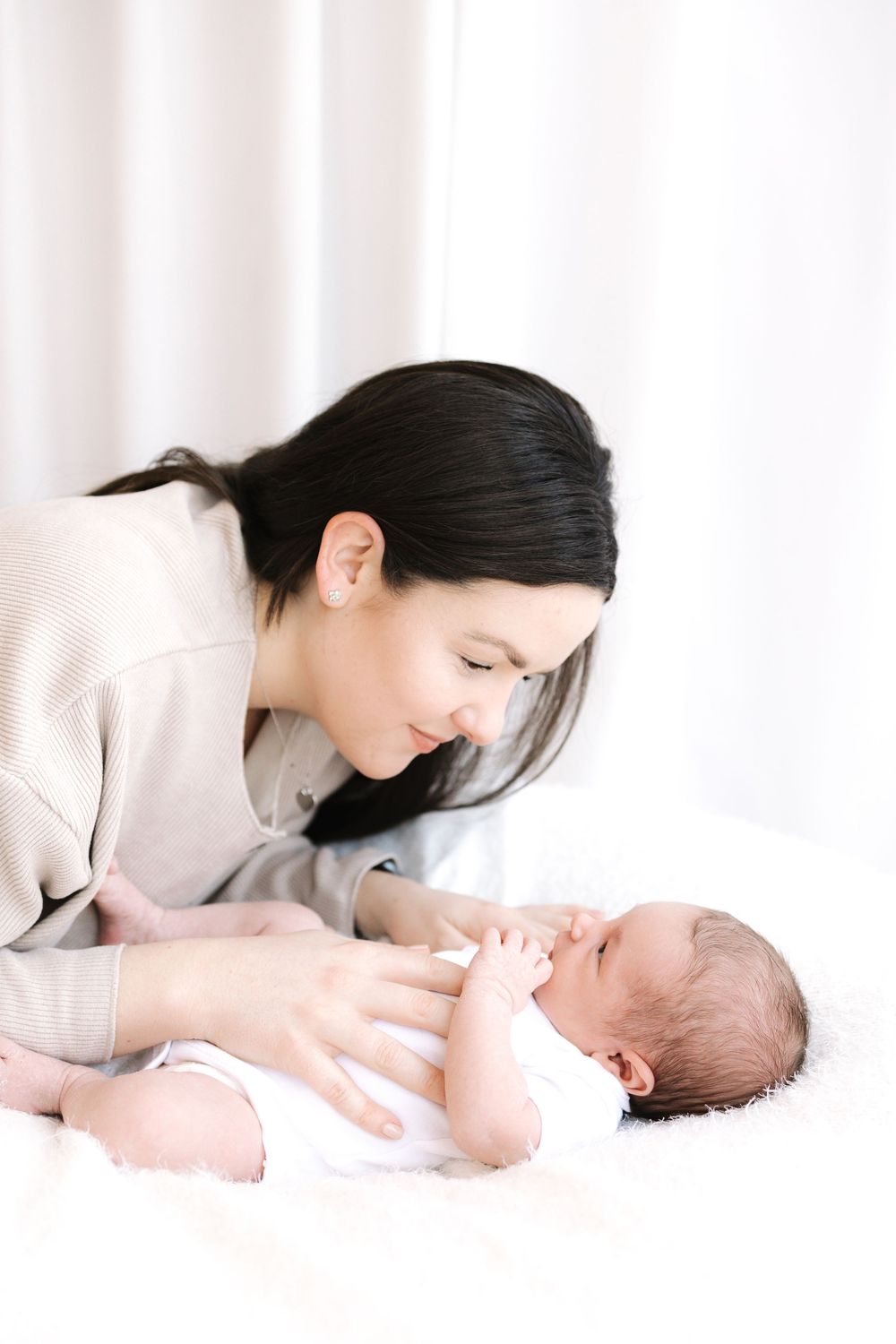 A young dark-haired mother tenderly gazes at her newborn baby lying on a white fluffy blanket.