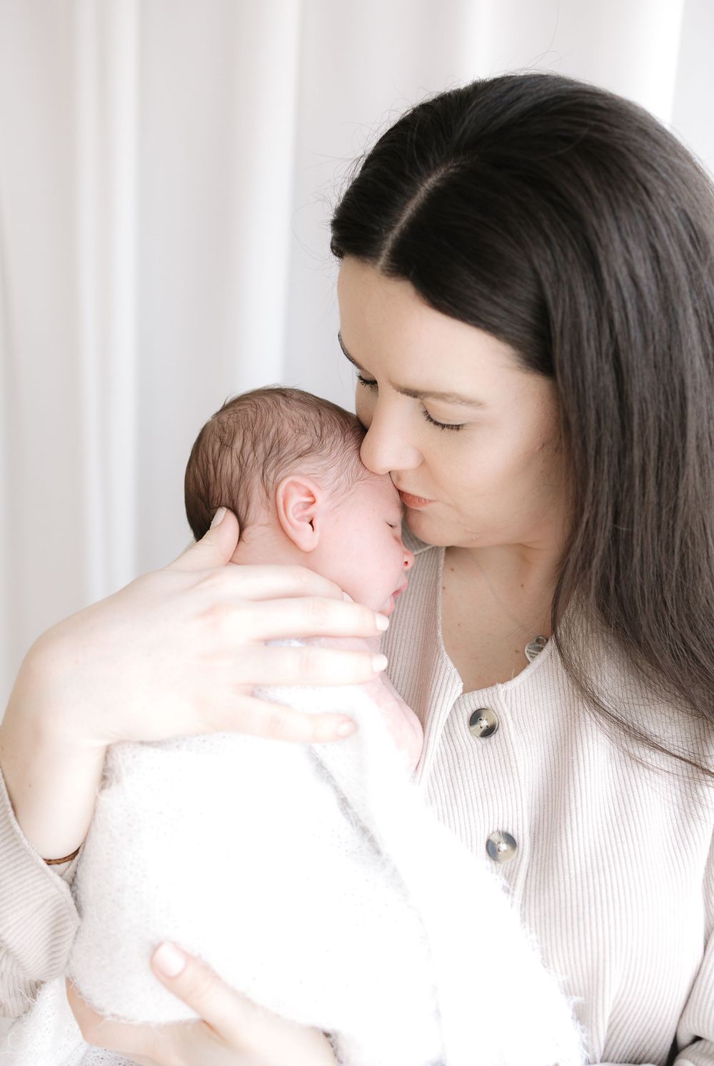 A mother with dark hair gently kisses her sleeping newborn baby wrapped in white, against a soft light background.