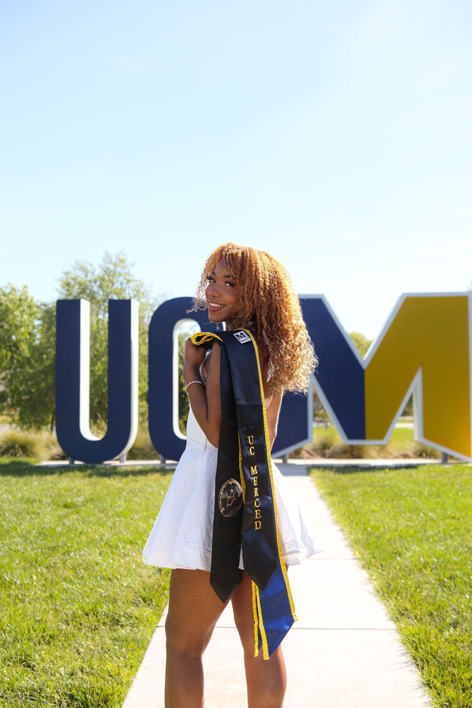 Smiling female graduate in white dress holds UC Merced graduation stole near large UCM campus sign.