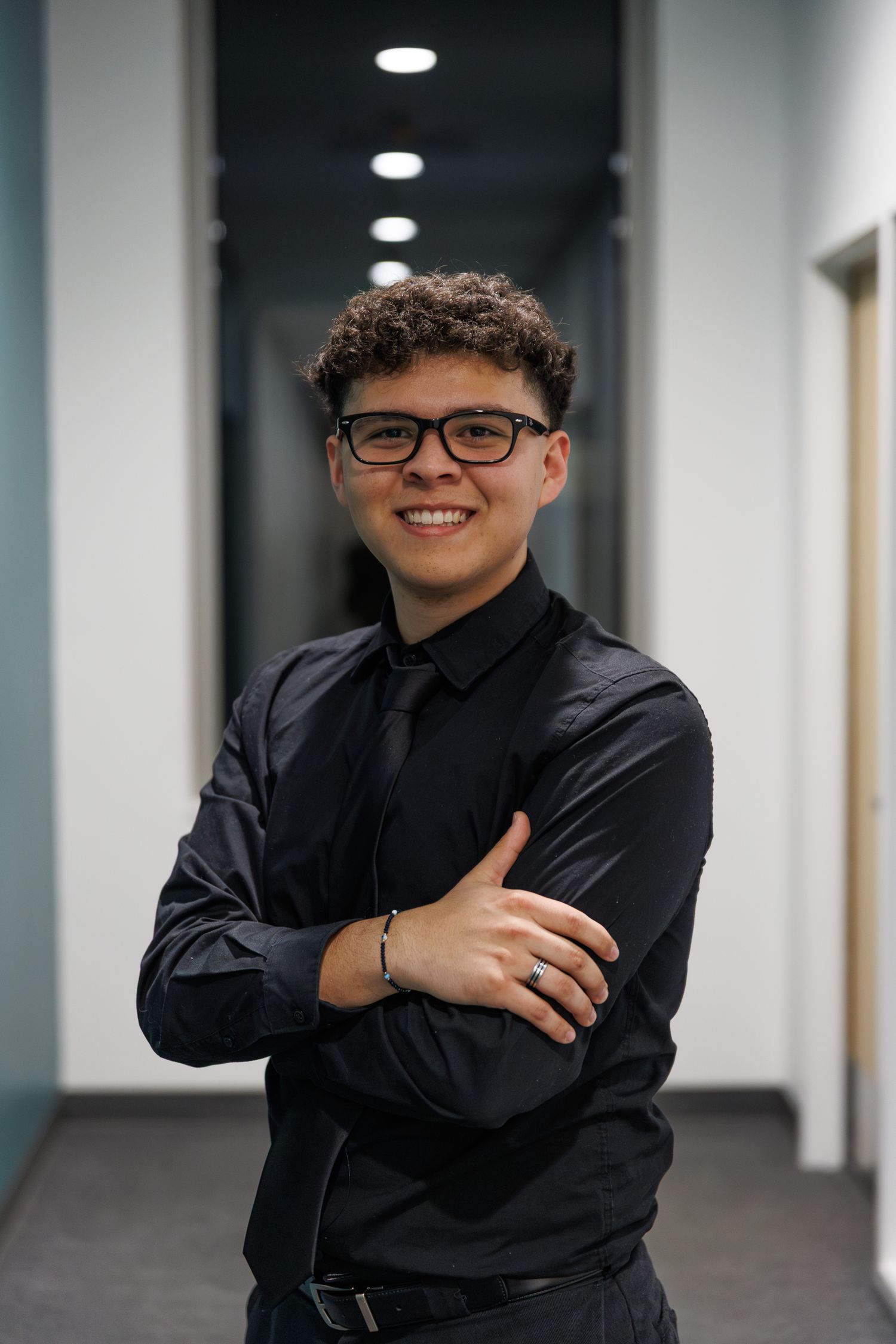 A young man with curly hair and glasses wearing a black shirt and tie smiling with arms crossed in an office hallway.