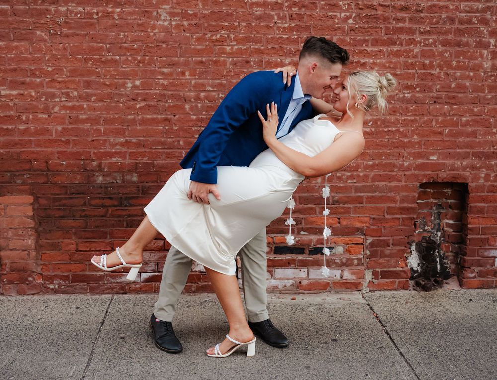 A romantic couple in formal attire dances and dips against a rustic red brick wall background.