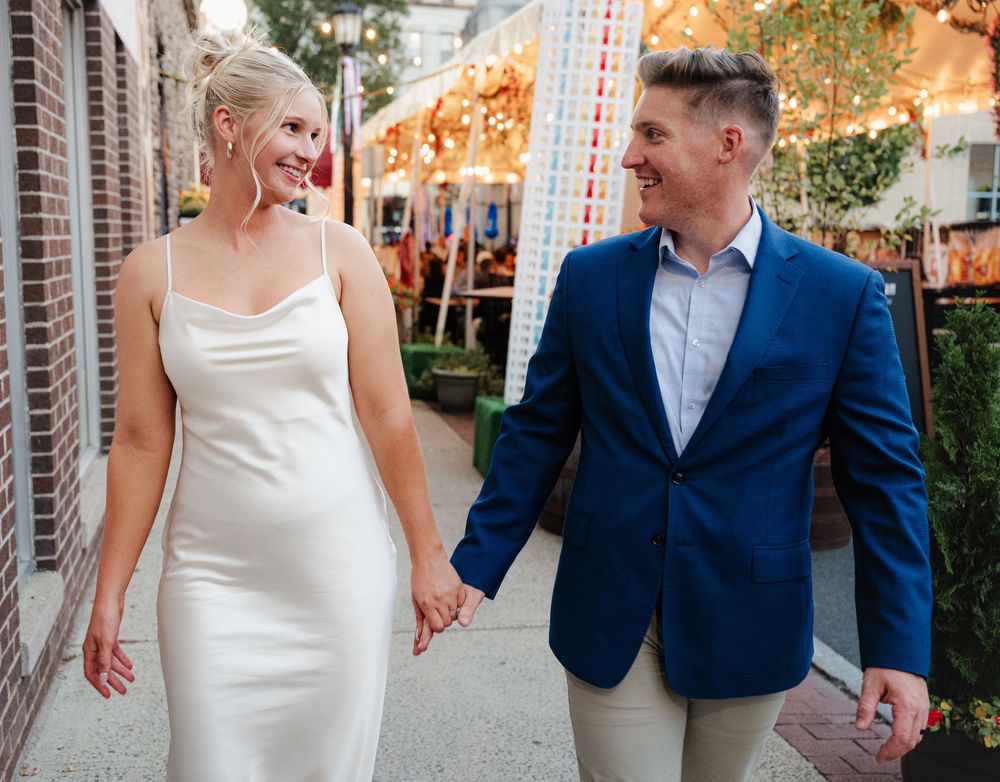 A couple in wedding attire walks hand in hand through a garden setting with brick walls and string lights overhead.