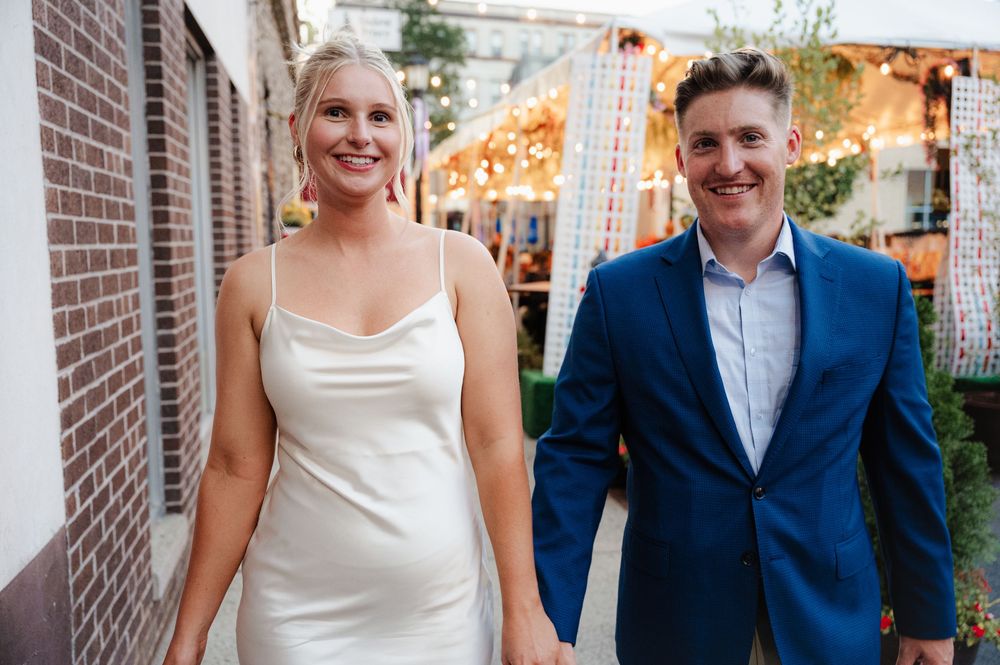 A couple in wedding attire stands together in a brick-lined walkway during an outdoor celebration.