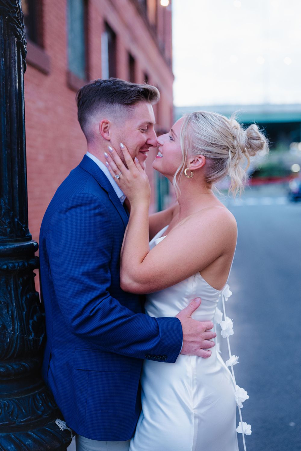 A couple in wedding attire share an intimate moment against a brick building backdrop.