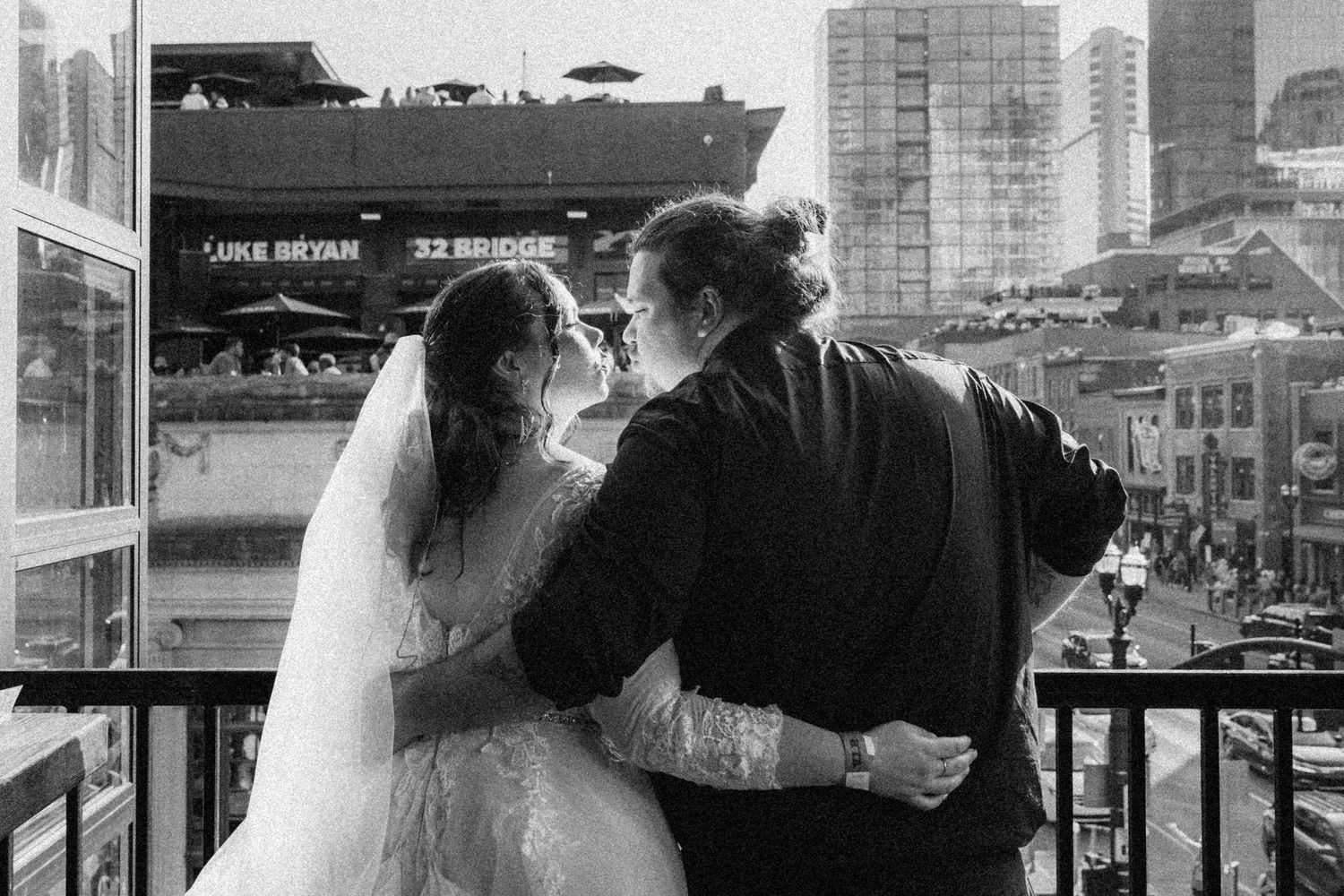 Newlyweds embrace while looking out at a cityscape with a cargo ship visible in the background.