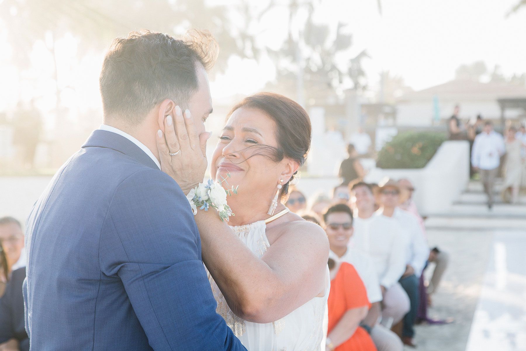 Emotional mother embraces groom during outdoor beach wedding ceremony.