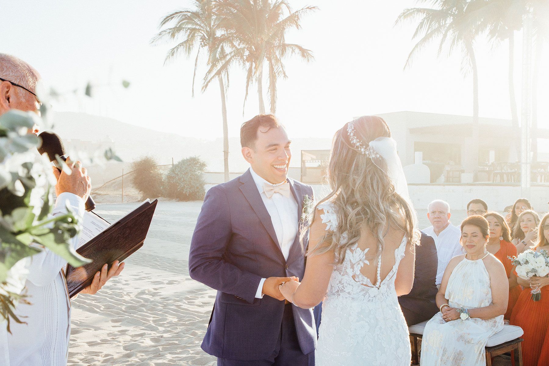 Groom smiles at bride during sunny beach wedding ceremony with palm trees.