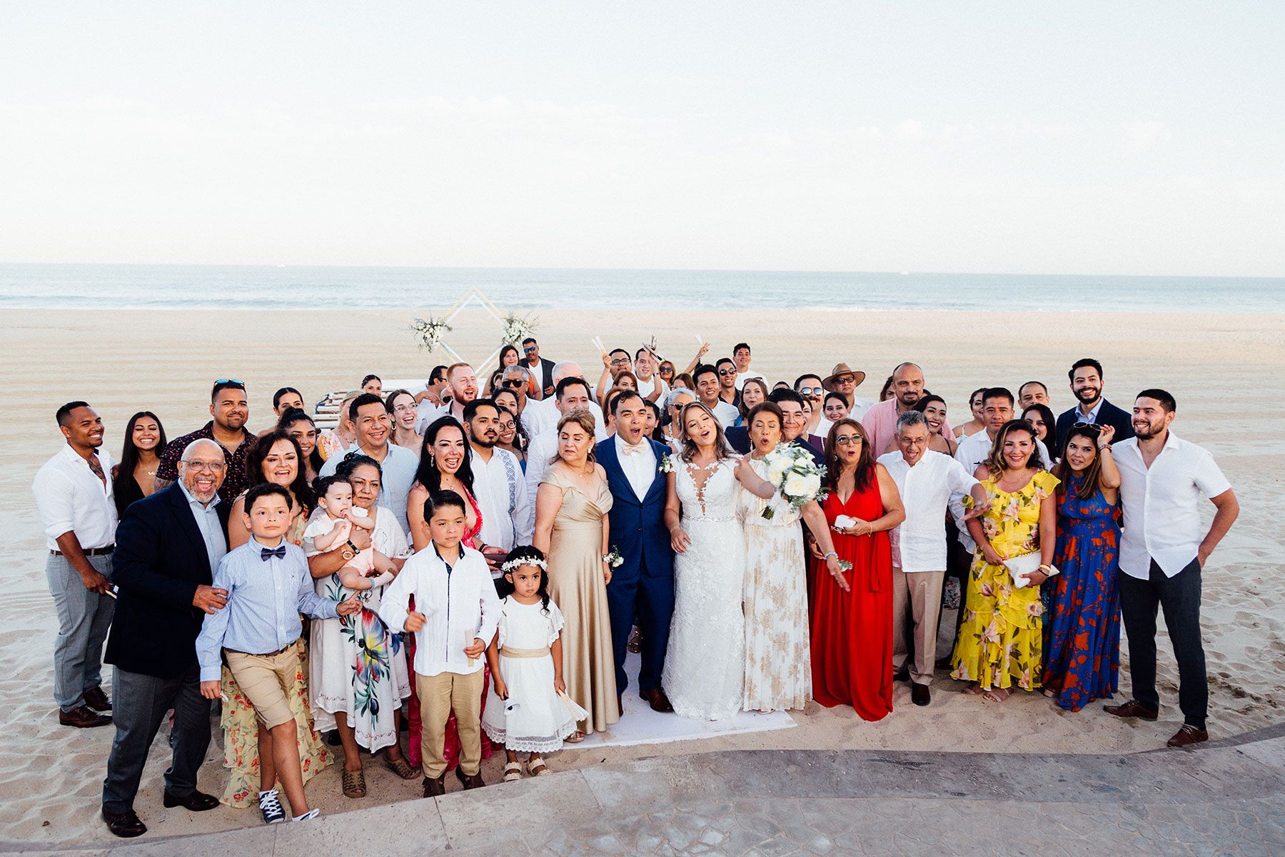 Large wedding party group photo on beach at sunset with ocean backdrop.