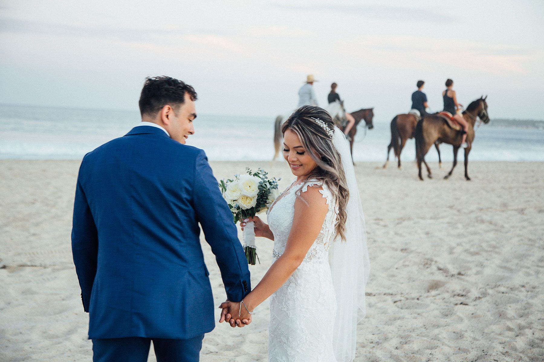 Newlyweds hold hands on beach with horses and riders in background.