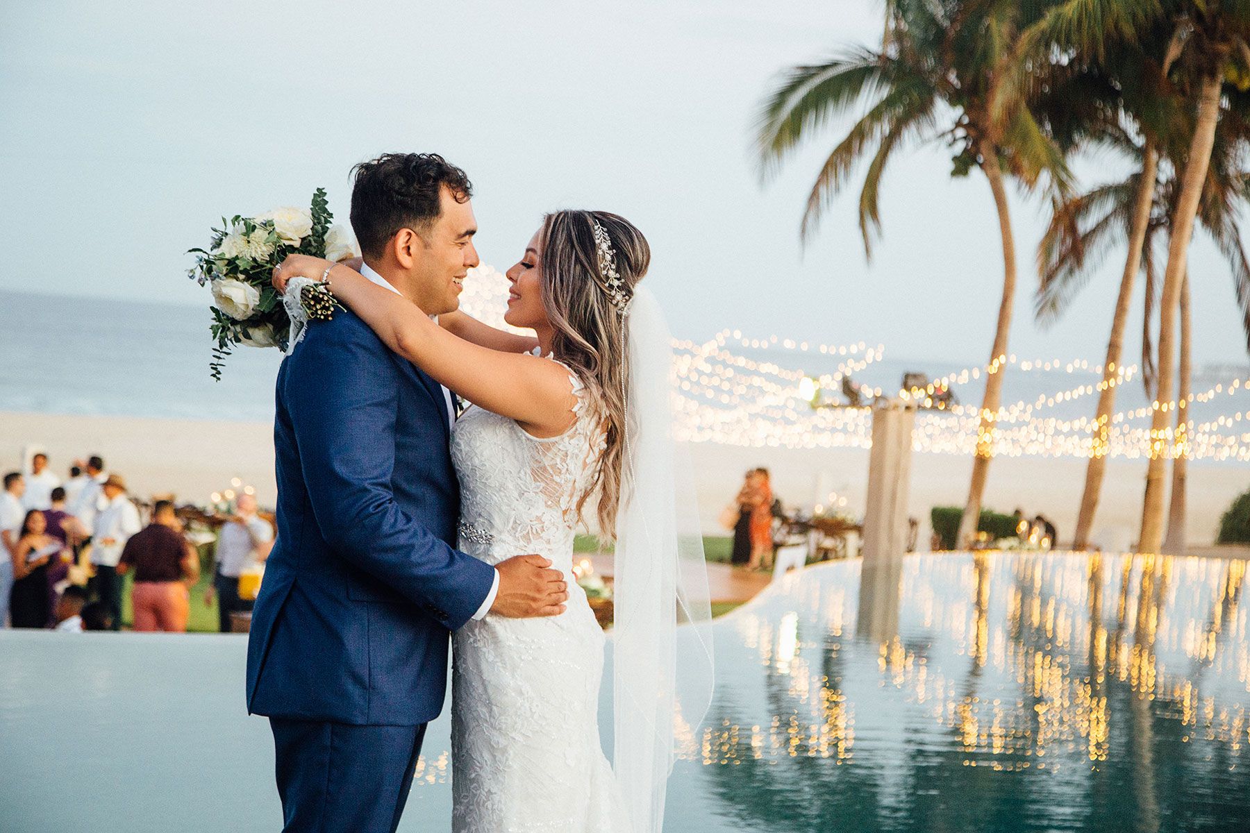 Bride and groom embrace romantically by infinity pool at twilight reception.