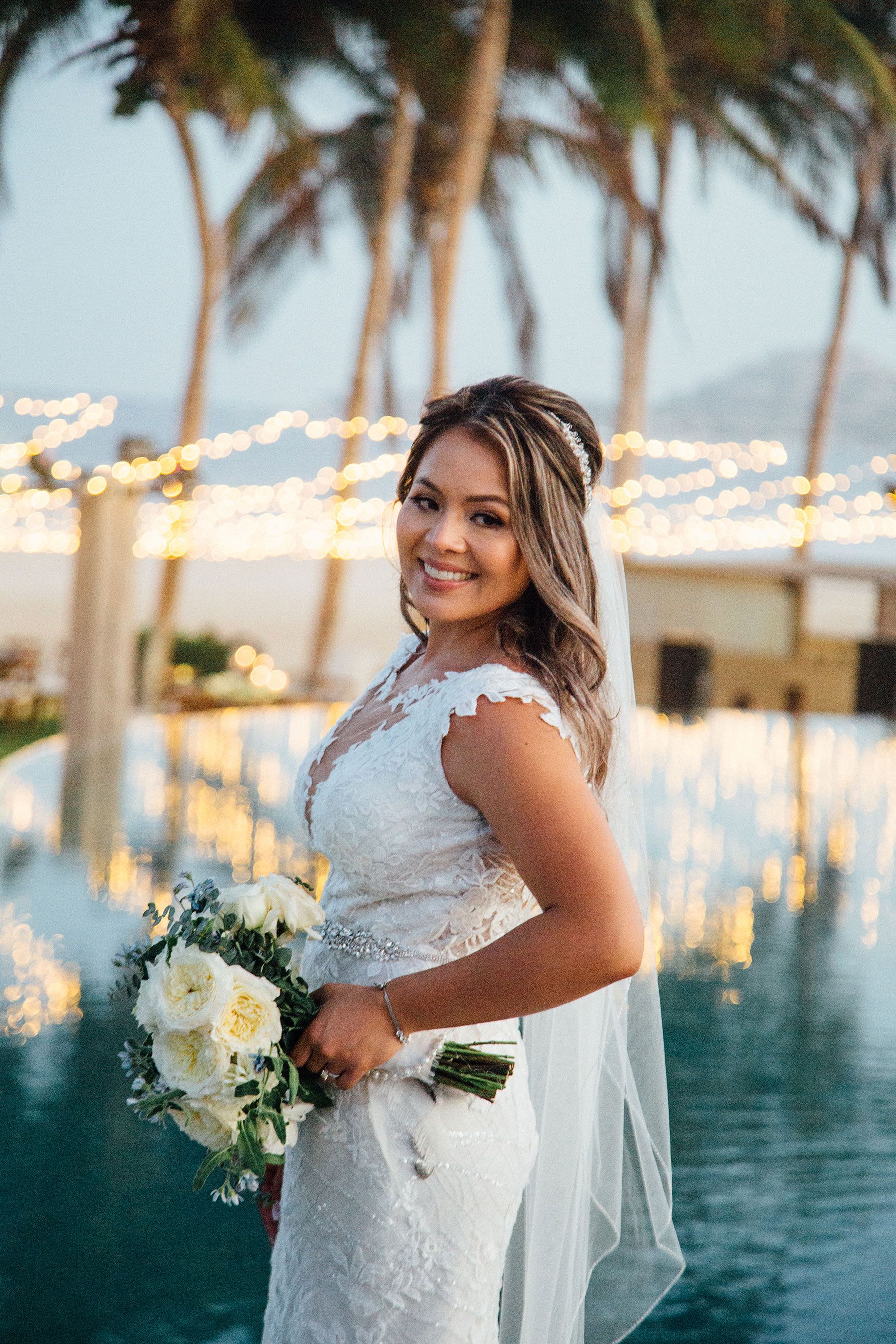 Smiling bride in lace gown holds white bouquet near palm trees and pool.