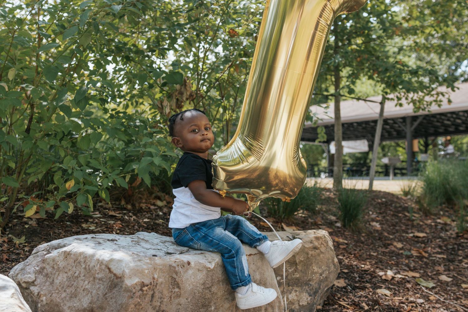 A child sits on a rock with a large gold number one balloon in a garden setting.