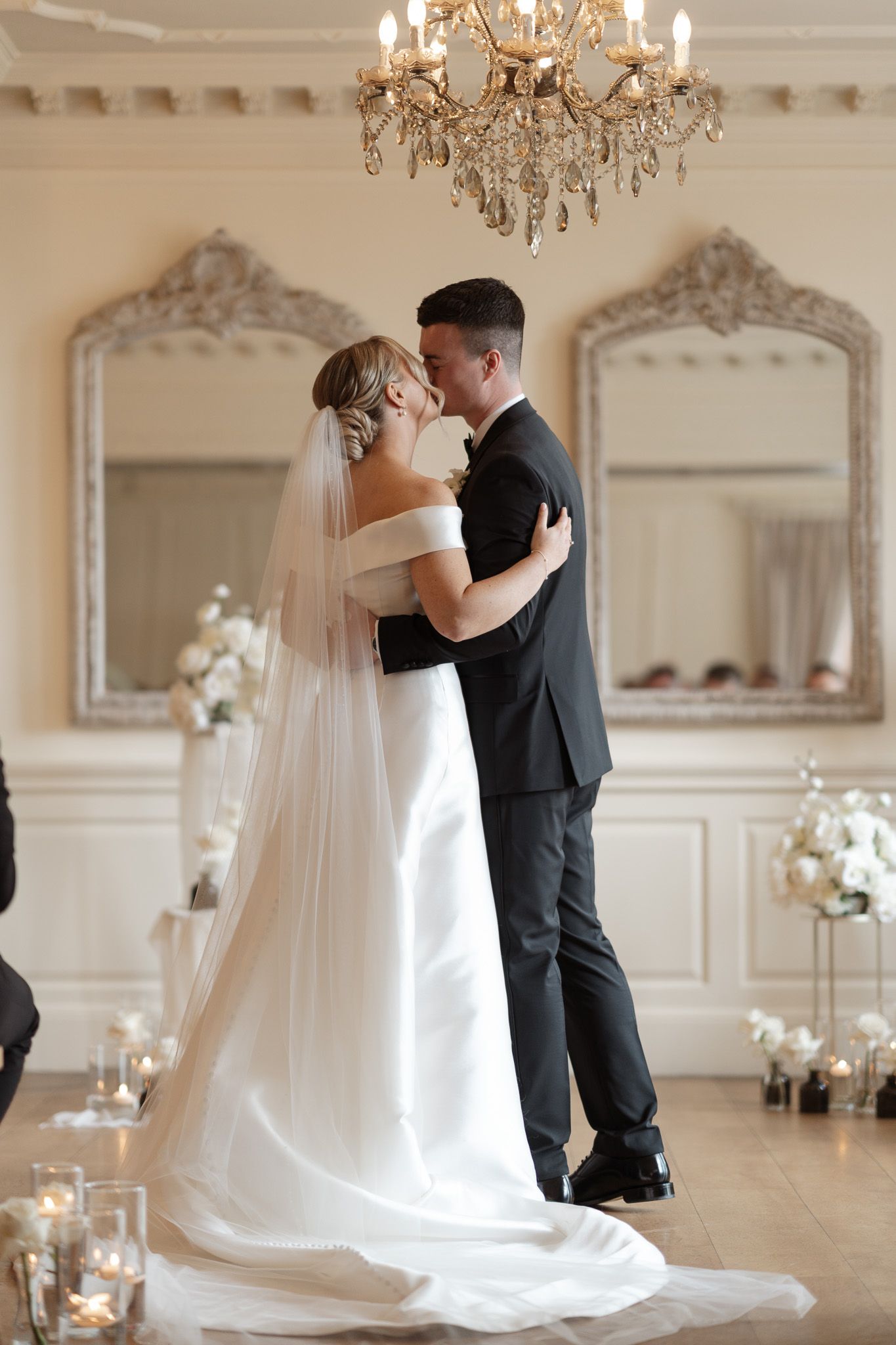 An elegant wedding couple embracing under a crystal chandelier in a room with ornate mirrors.