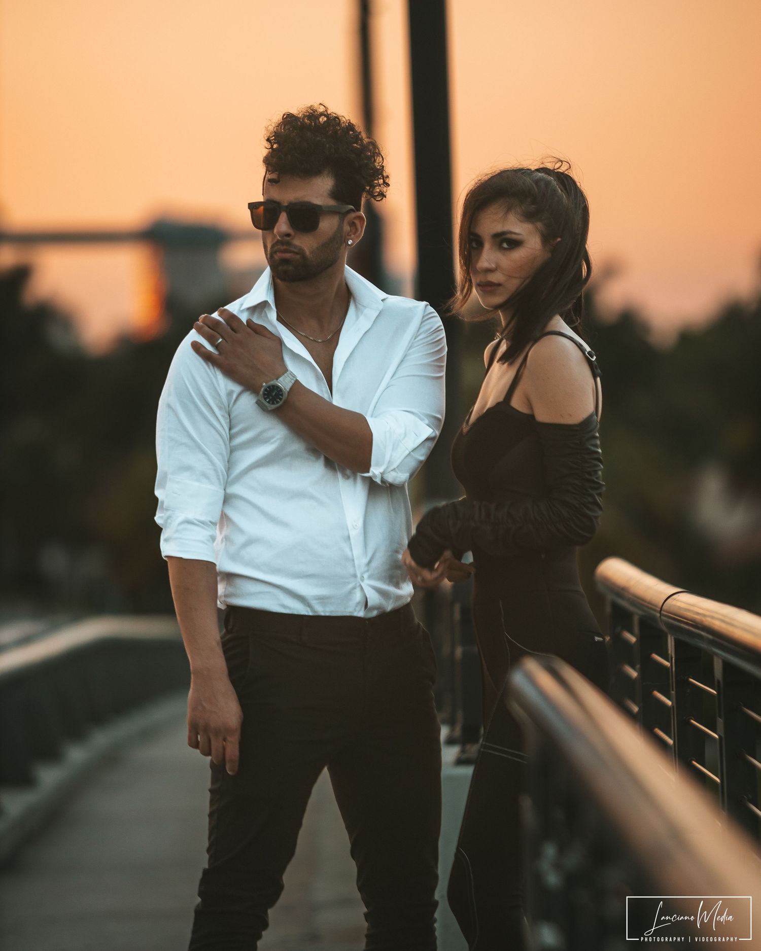 Stylish couple in formal attire posing together at sunset on a bridge walkway.