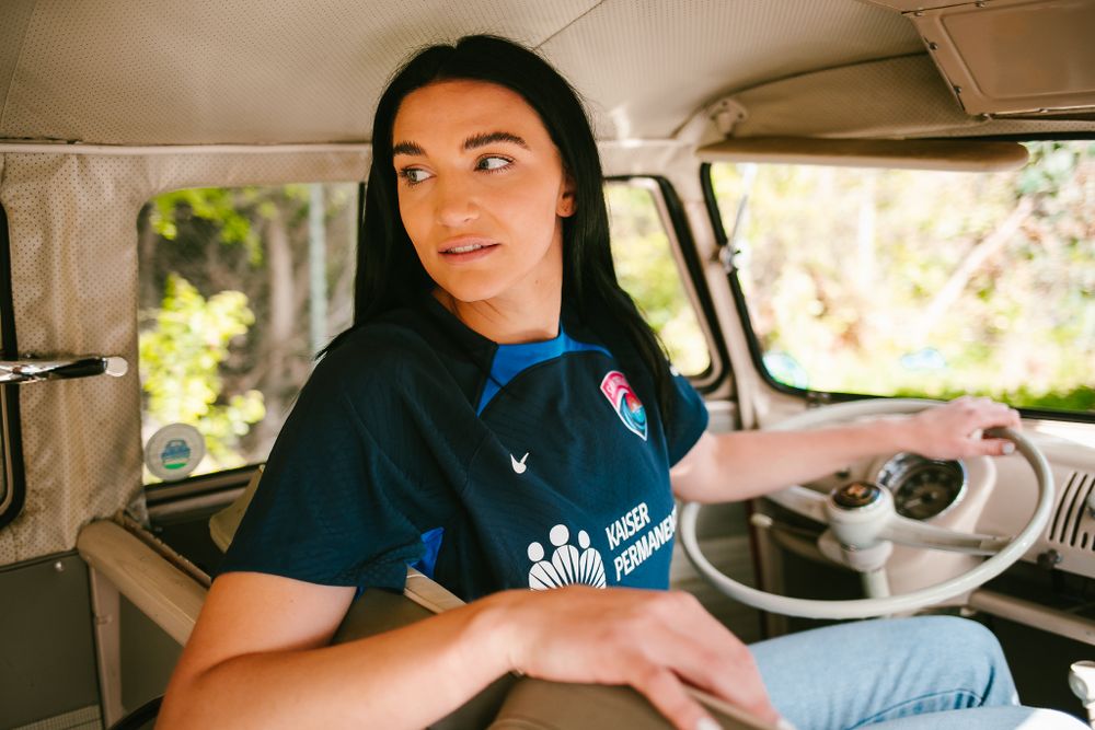 Dark-haired female soccer player in blue Kaiser Permanente jersey sits in vintage van driver seat.
