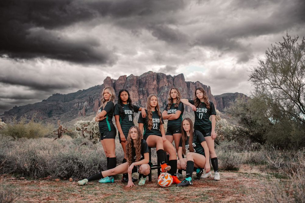 Girls soccer team poses with equipment in front of the rugged Superstition Mountains under dramatic storm clouds.