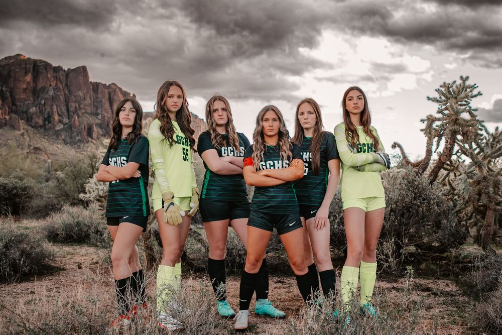 Six female soccer players pose confidently in green uniforms against the scenic Arizona desert mountain backdrop.